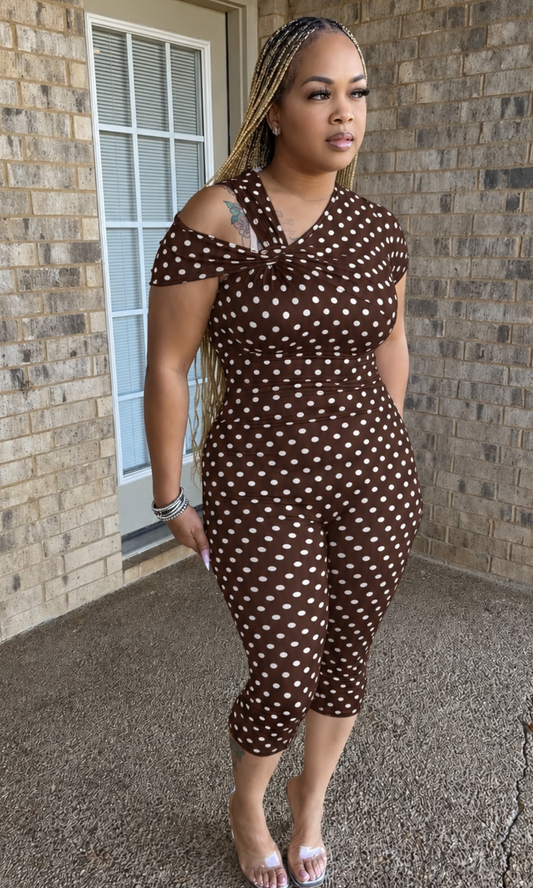 Woman modeling the Brown Polka Babe Jumpsuit featuring a chocolate brown and white polka dot print, twisted asymmetrical neckline, off-shoulder detail, and fitted capri-length legs.