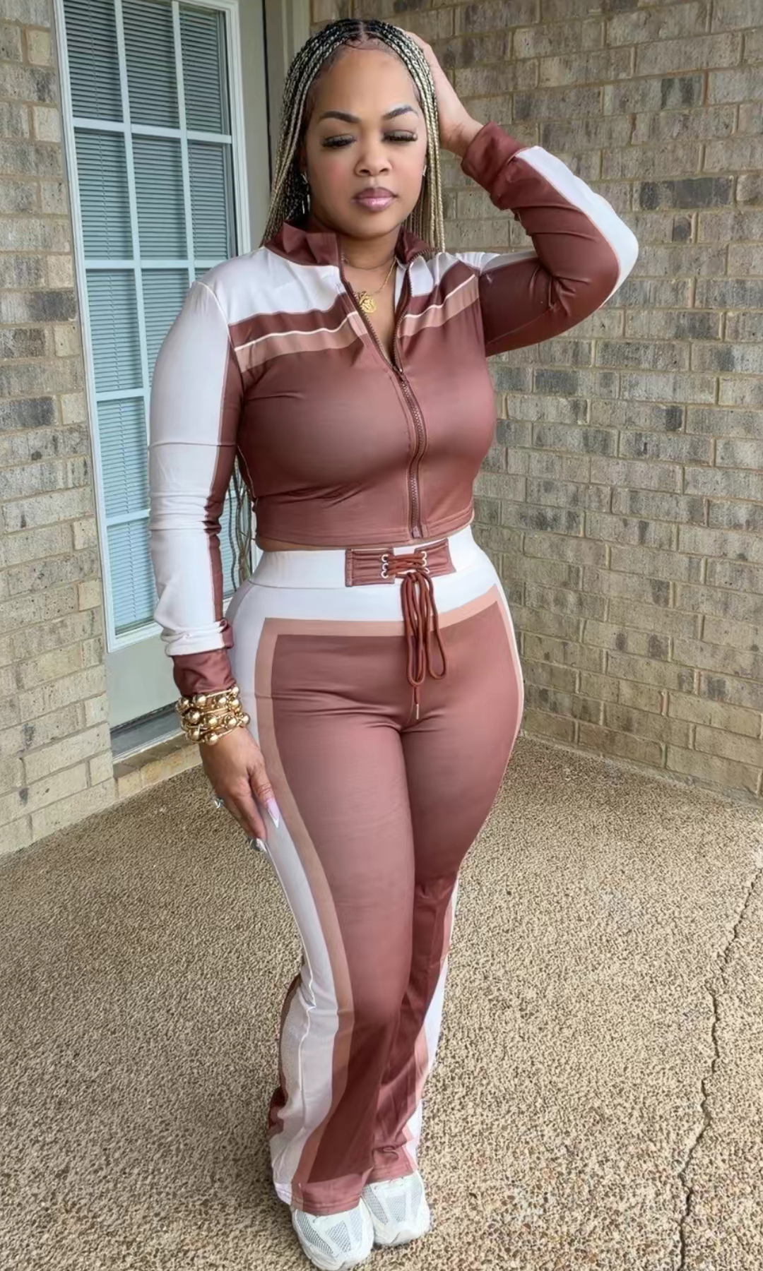 A woman modeling a matching two-piece tracksuit with a zip-up long-sleeve top and high-waisted flared pants in a dusty rose and white colorway, standing outdoors against a brick wall.