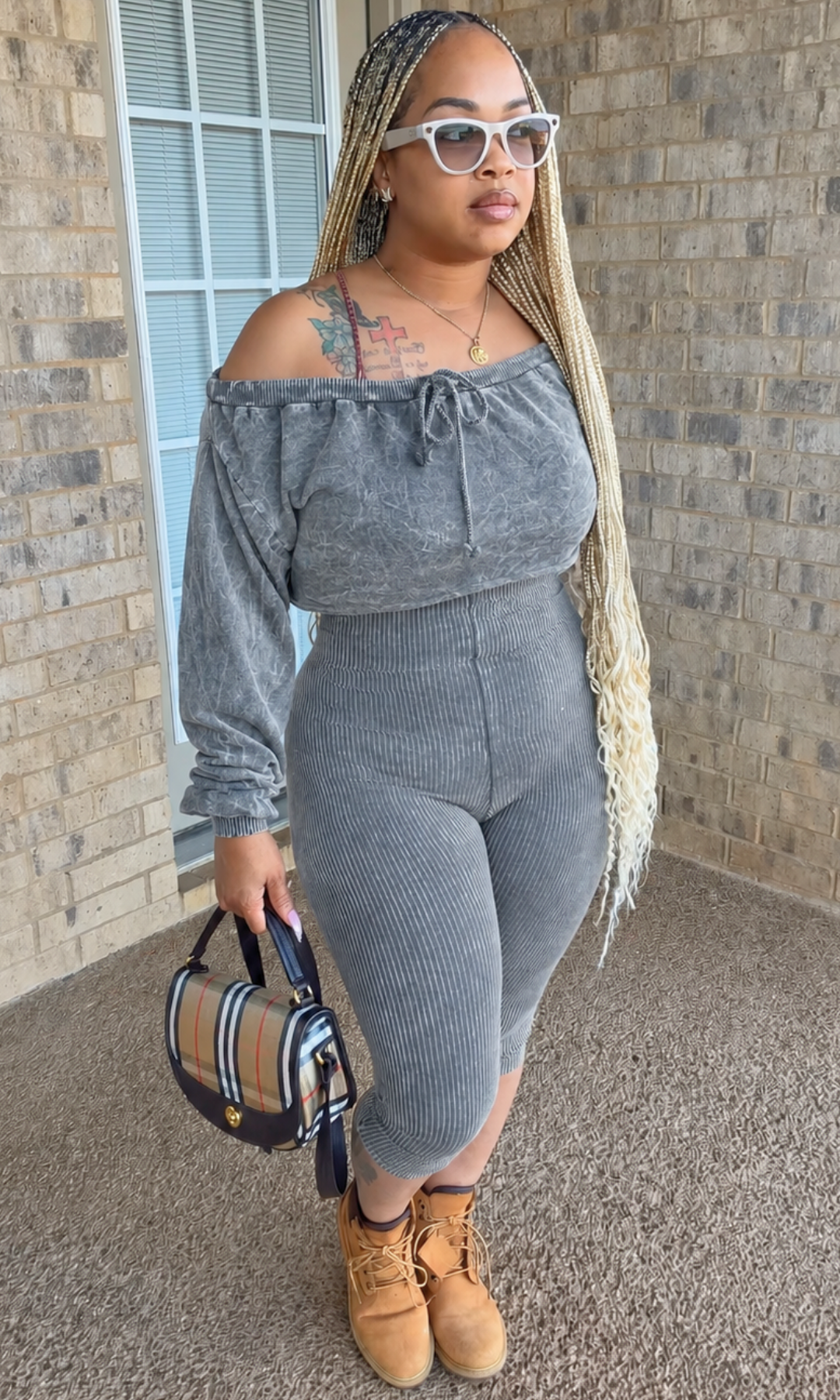 A woman modeling a gray ribbed off-the-shoulder long-sleeve crop top and matching ribbed leggings set, styled with white sunglasses, a plaid handbag, and tan lace-up boots, standing in front of a brick wall.