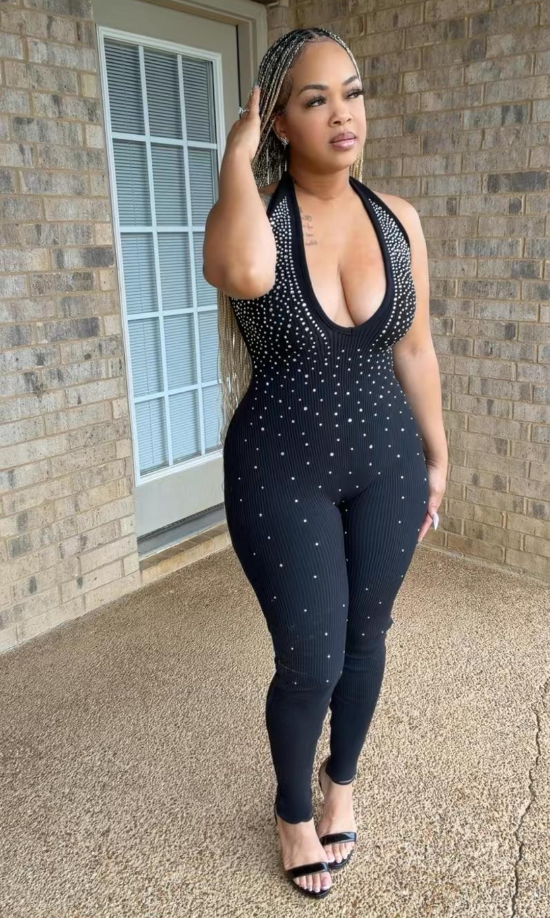 Woman modeling the Diamond Doll Jumpsuit, a fitted black ribbed one-piece with a plunging halter neckline, sparkling rhinestone embellishments, and full-length legs.