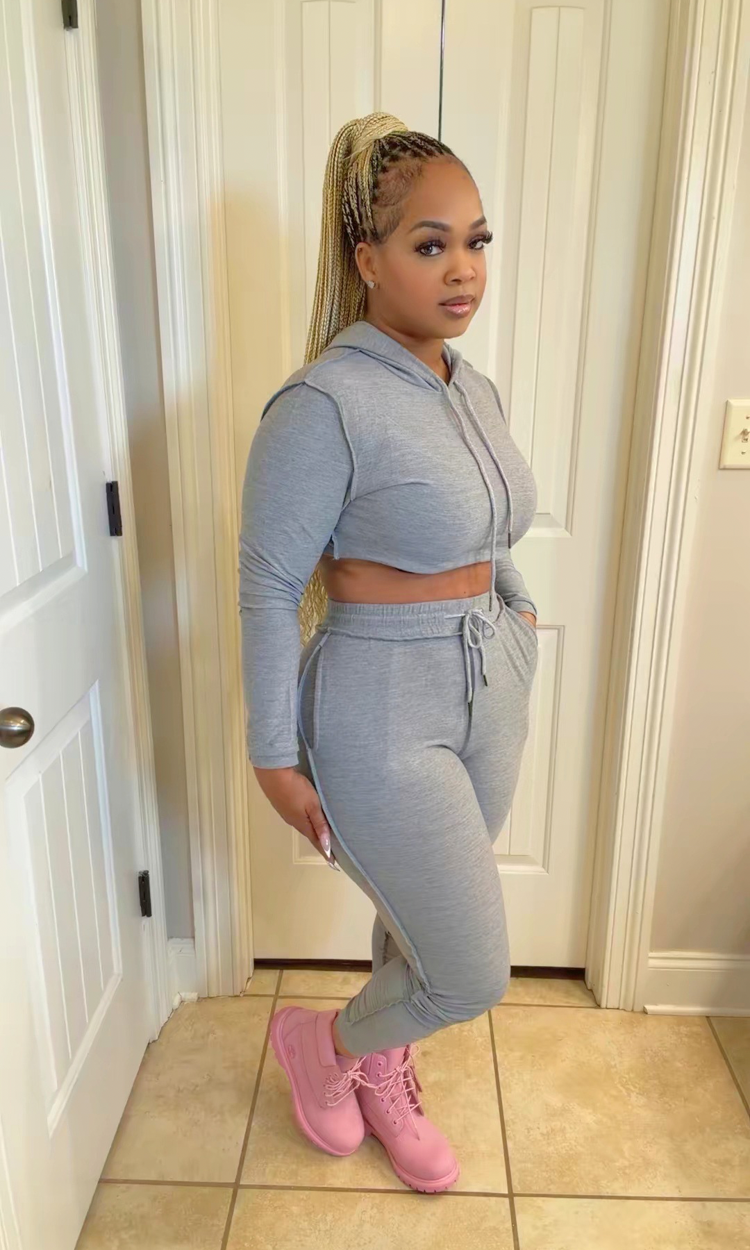 A woman modeling a light gray two-piece loungewear set consisting of a cropped zip-up hoodie and matching jogger pants, paired with pink sneakers, standing in front of white interior doors.