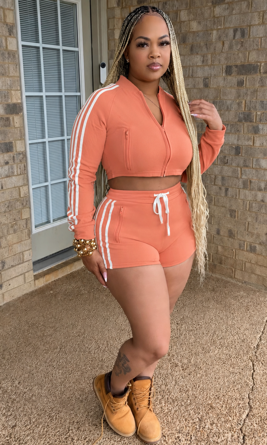 Woman modeling the Race and Track Peach Set featuring a peach cropped zip-up jacket with white racing stripes and coordinating high-waisted shorts with a white drawstring waistband.