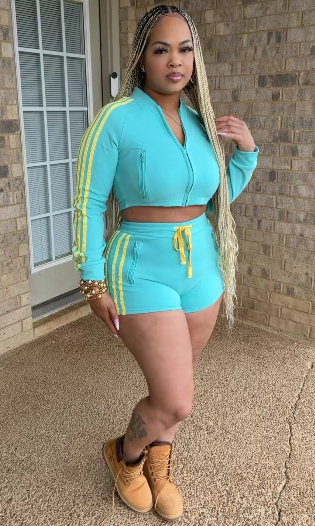 Woman modeling the Race and Track Set in turquoise blue with yellow racing stripes, featuring a long-sleeve cropped zip-up jacket and matching high-waisted fitted shorts.
