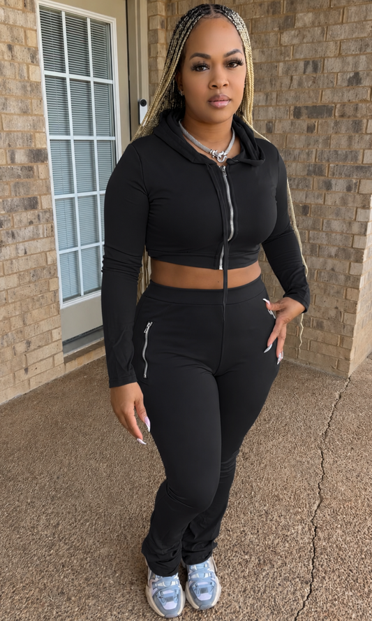 Woman modeling the Shadow Hoodie Set featuring a black cropped zip-up hoodie with drawstrings, long sleeves, and coordinating high-waisted fitted pants with silver zipper pocket details.