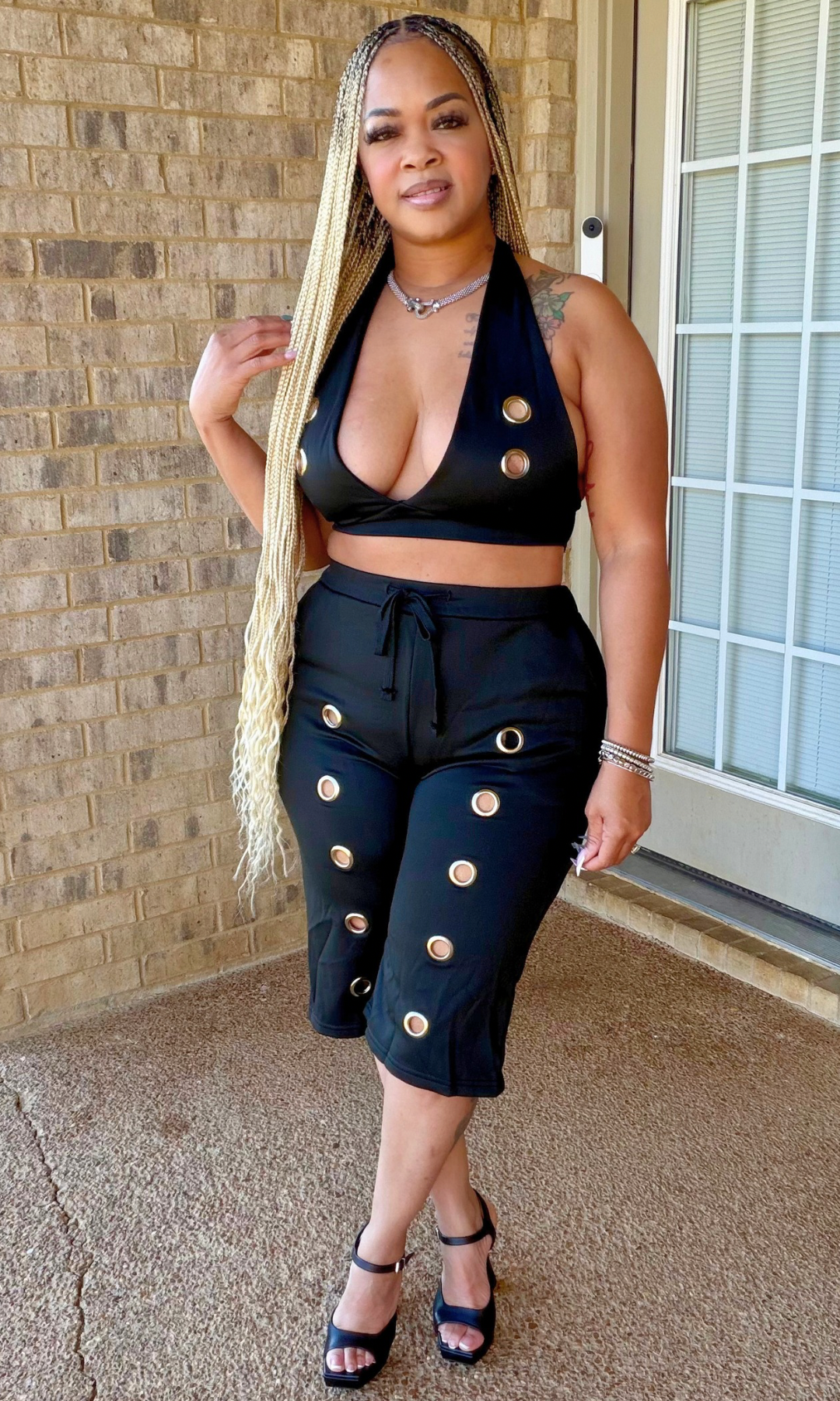 Woman modeling the black Wrestler Set featuring a plunging halter crop top with gold-tone grommet accents and matching high-waisted knee-length shorts, front view.